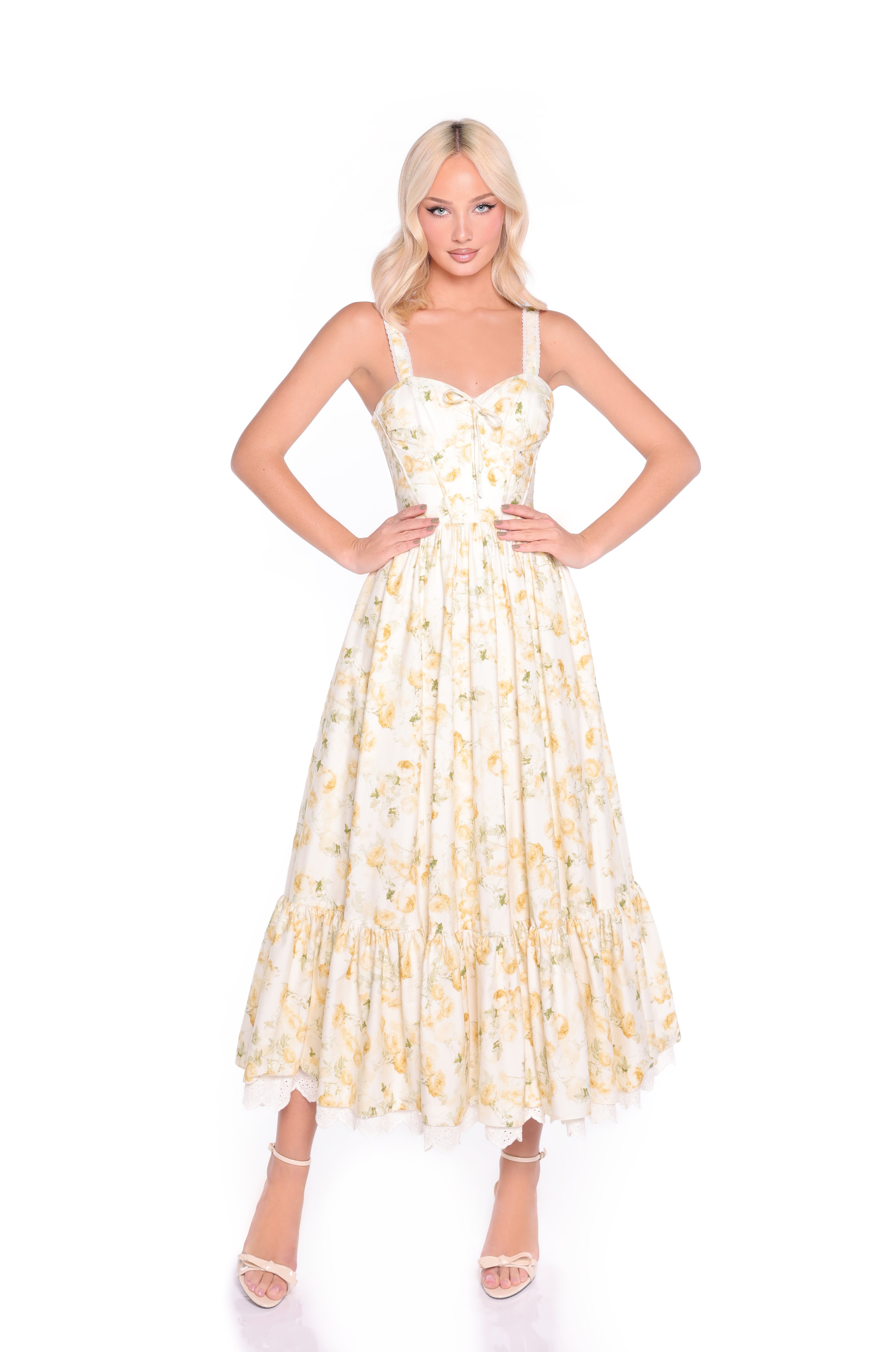 A woman stands hands on hips against a plain white background, modeling the Buttercream Floral Casual Midi Dress: Marigold, a sleeveless white dress with yellow floral print, corset bodice, and midi length.
